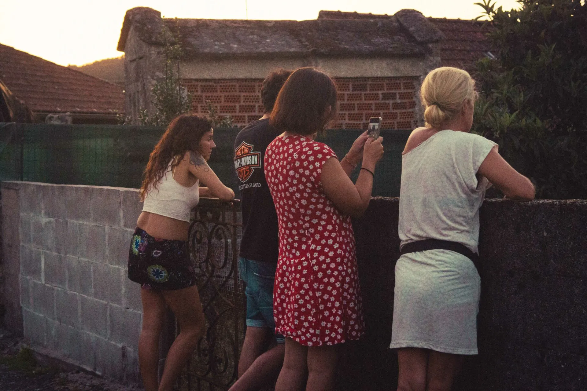 Three people seen from behind leaning on a low stone wall at sunset, one taking a photo with a phone, in front of an old tiled-roof house