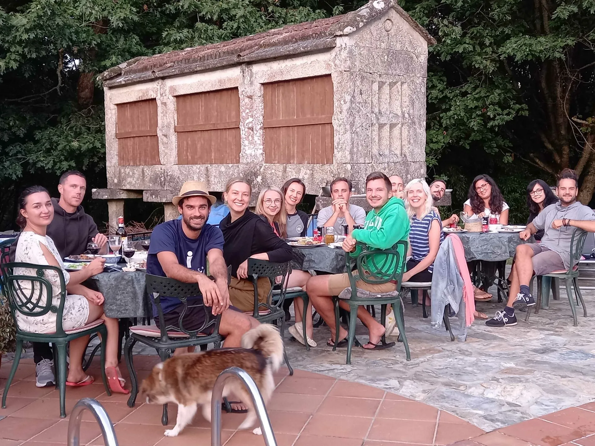 Around fifteen people having dinner at long outdoor tables in front of a stone hórreo, with a dog walking past