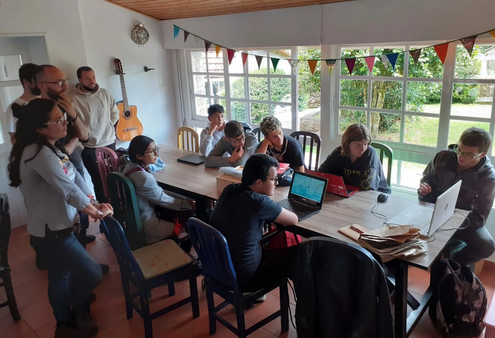 People working on laptops and papers around a long table in a bright room with bunting and a guitar on the wall