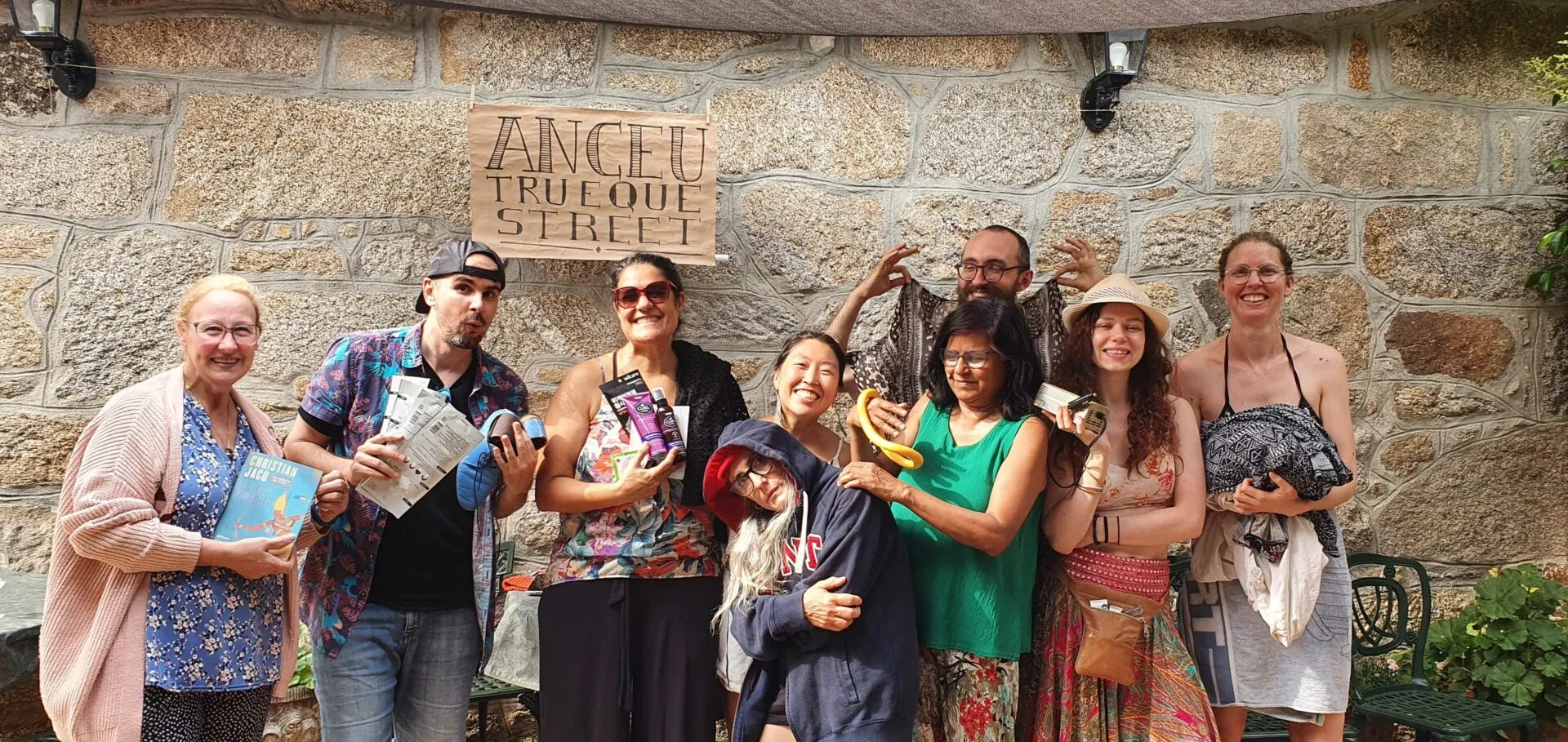 Nine cheerful people in colourful clothes posing against a stone wall under a wooden sign, holding flyers and small objects
