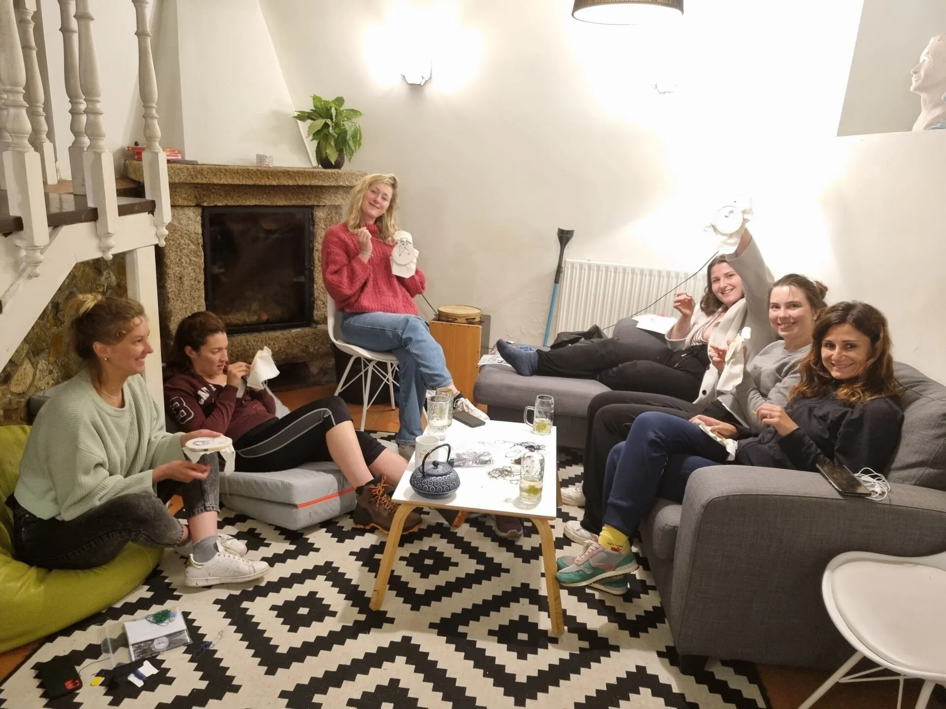 Six women relaxing on sofas and cushions around a stone fireplace, holding embroidery hoops in a cosy living room