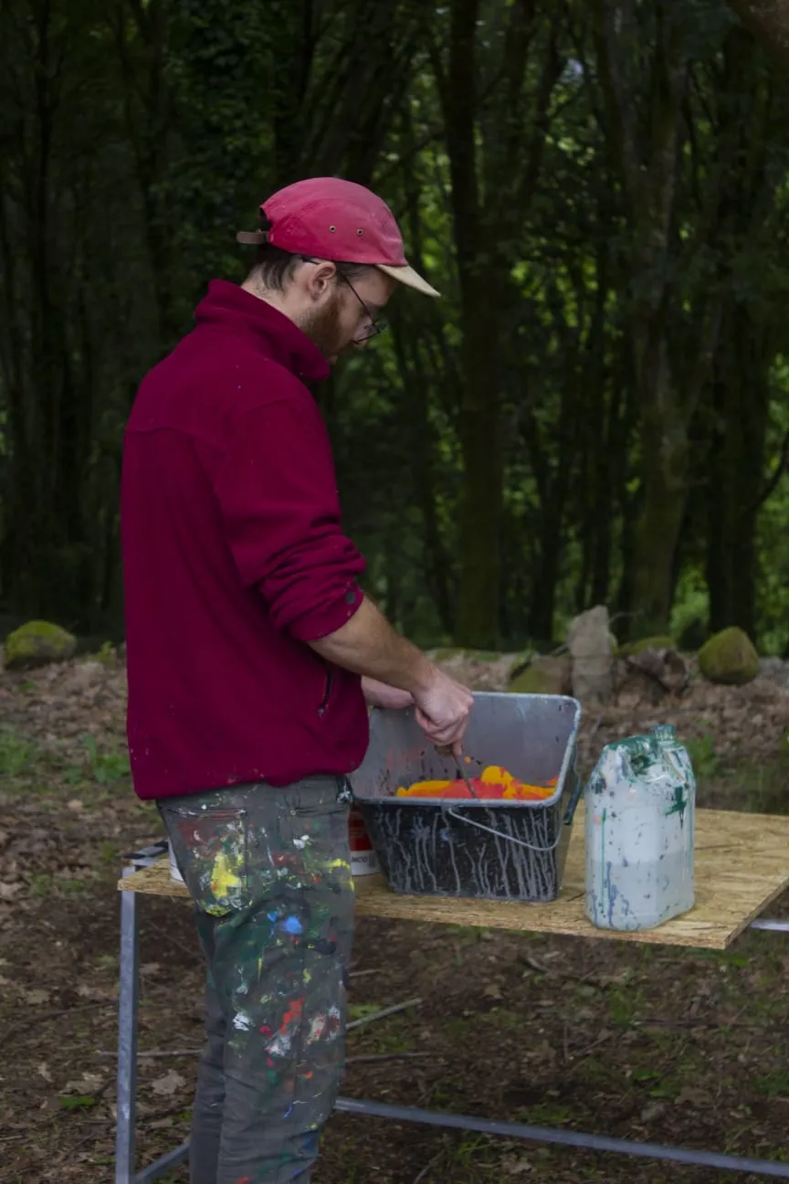 Man in a red cap and paint-spattered trousers stirring orange paint in a bucket on a trestle table in the woods