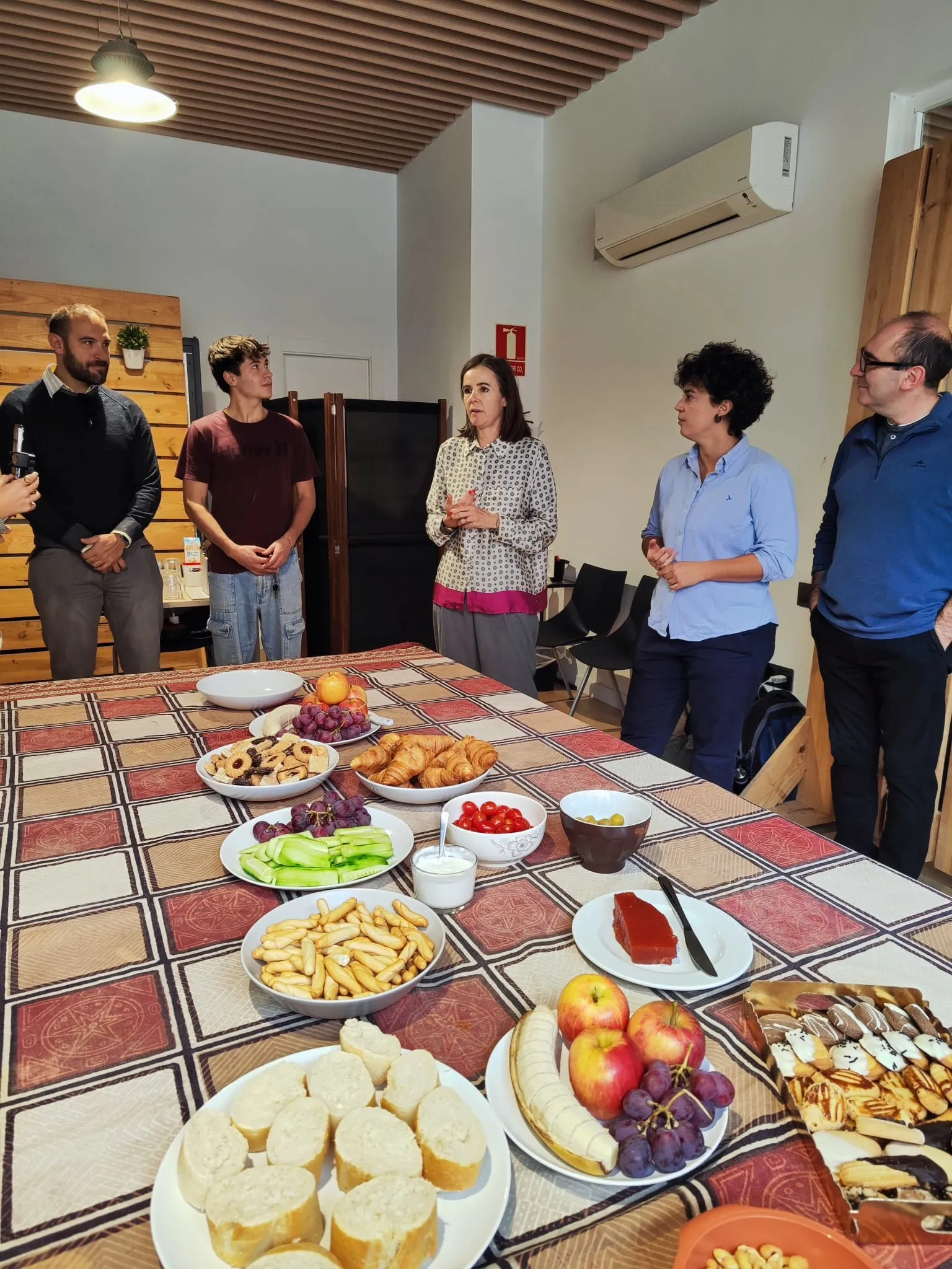 Five people standing around a long table laid with fruit, croissants, breadsticks and pastries as a woman speaks