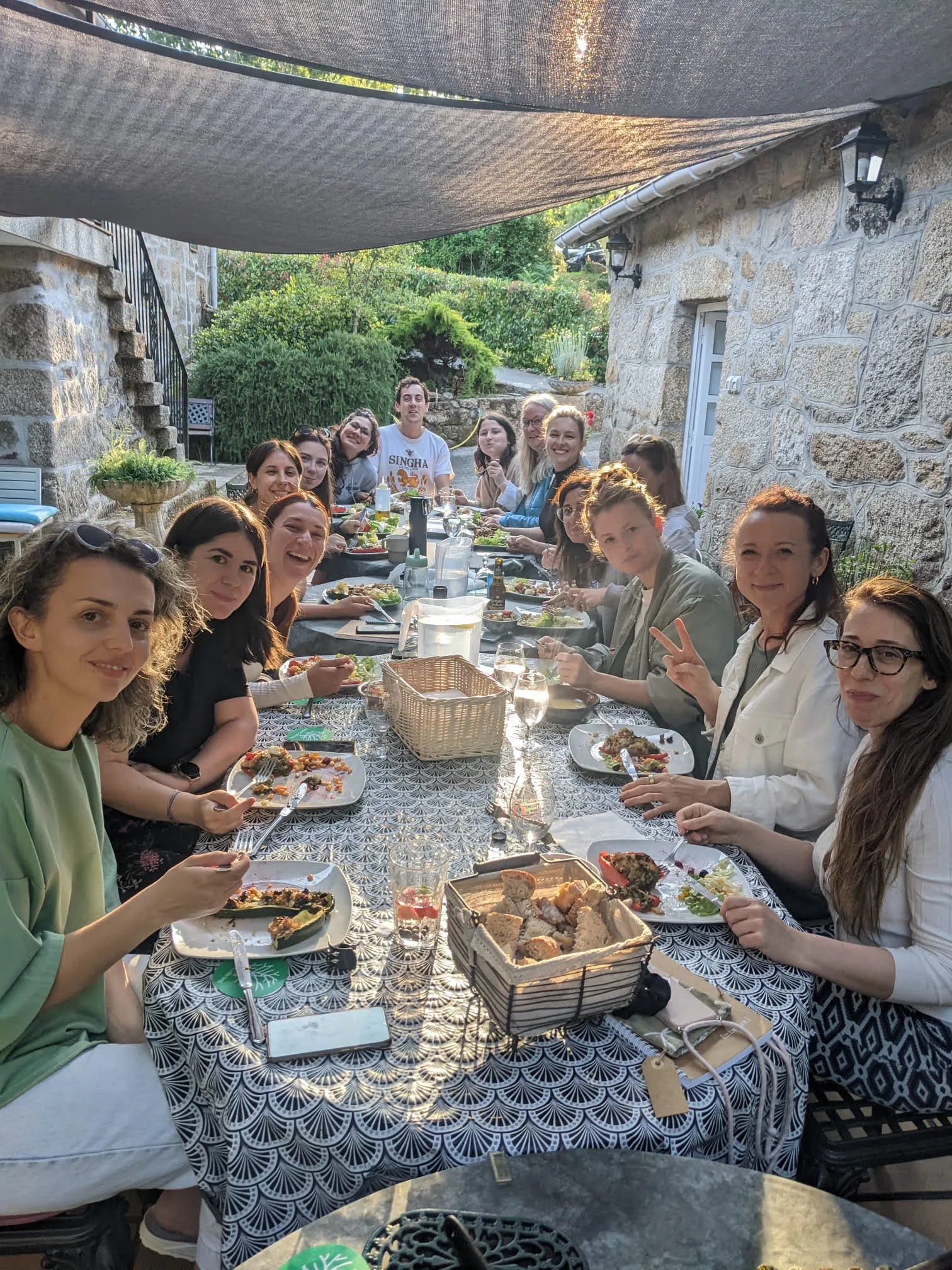 Long table of mostly women eating stuffed vegetables under a shade sail beside a stone house