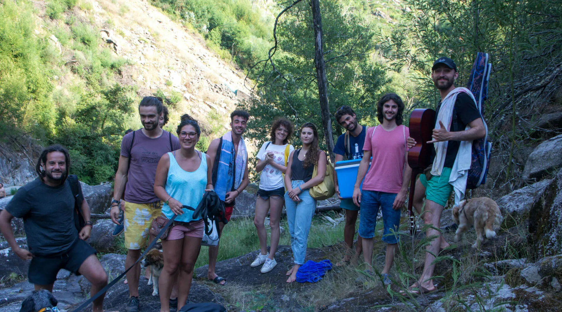 Ten people with towels, a guitar and a cooler bag posing beside a rocky stream in a wooded gorge, with a dog at the right