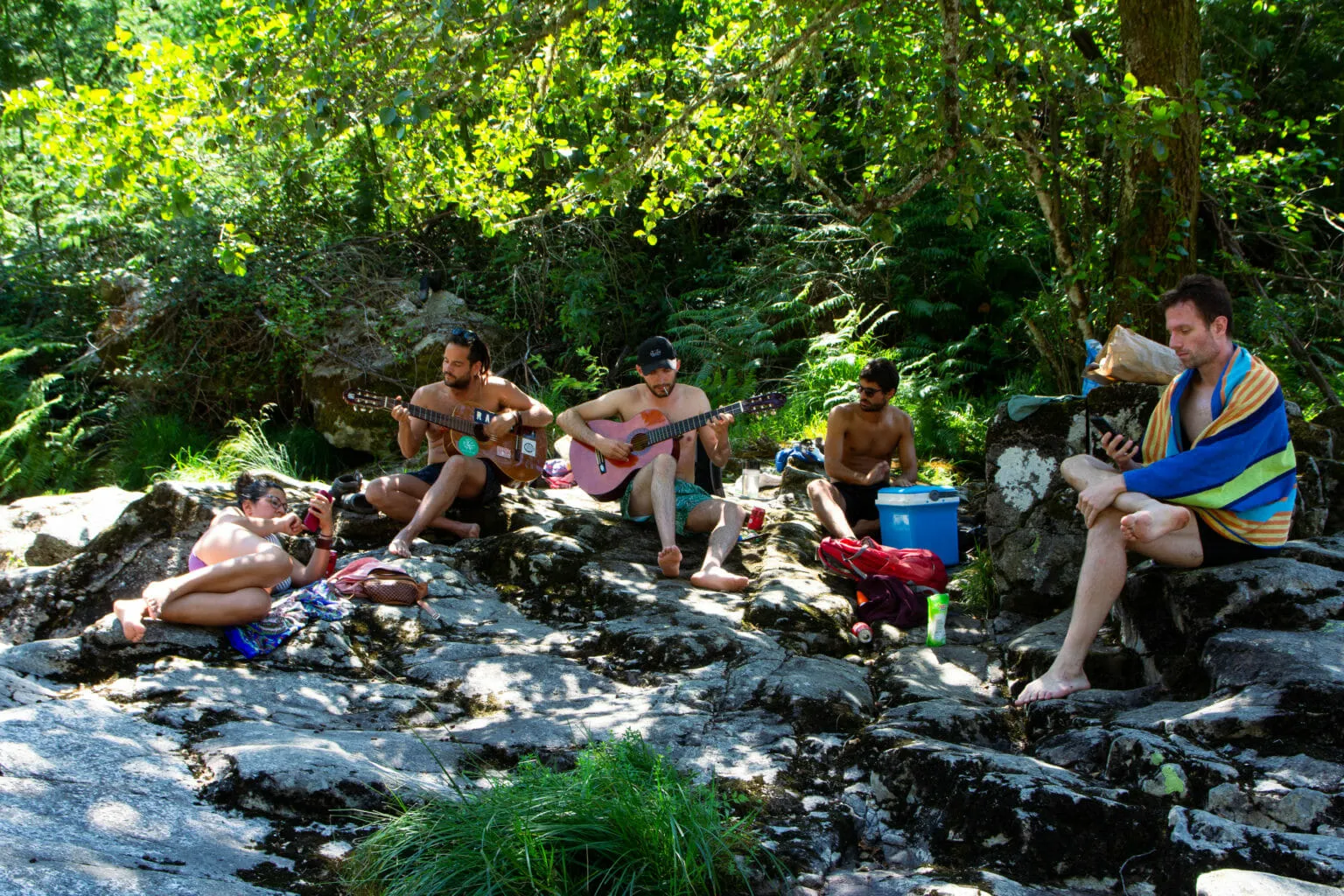 People relaxing on sunlit rocks beside a stream, two of them playing guitars