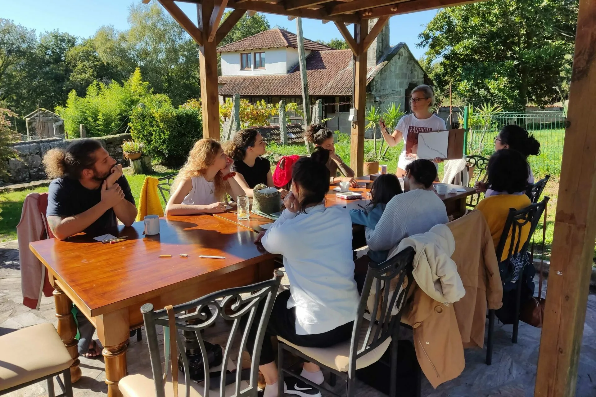 About ten people at a long wooden table under a pergola while a woman standing shows them a drawing, village houses behind