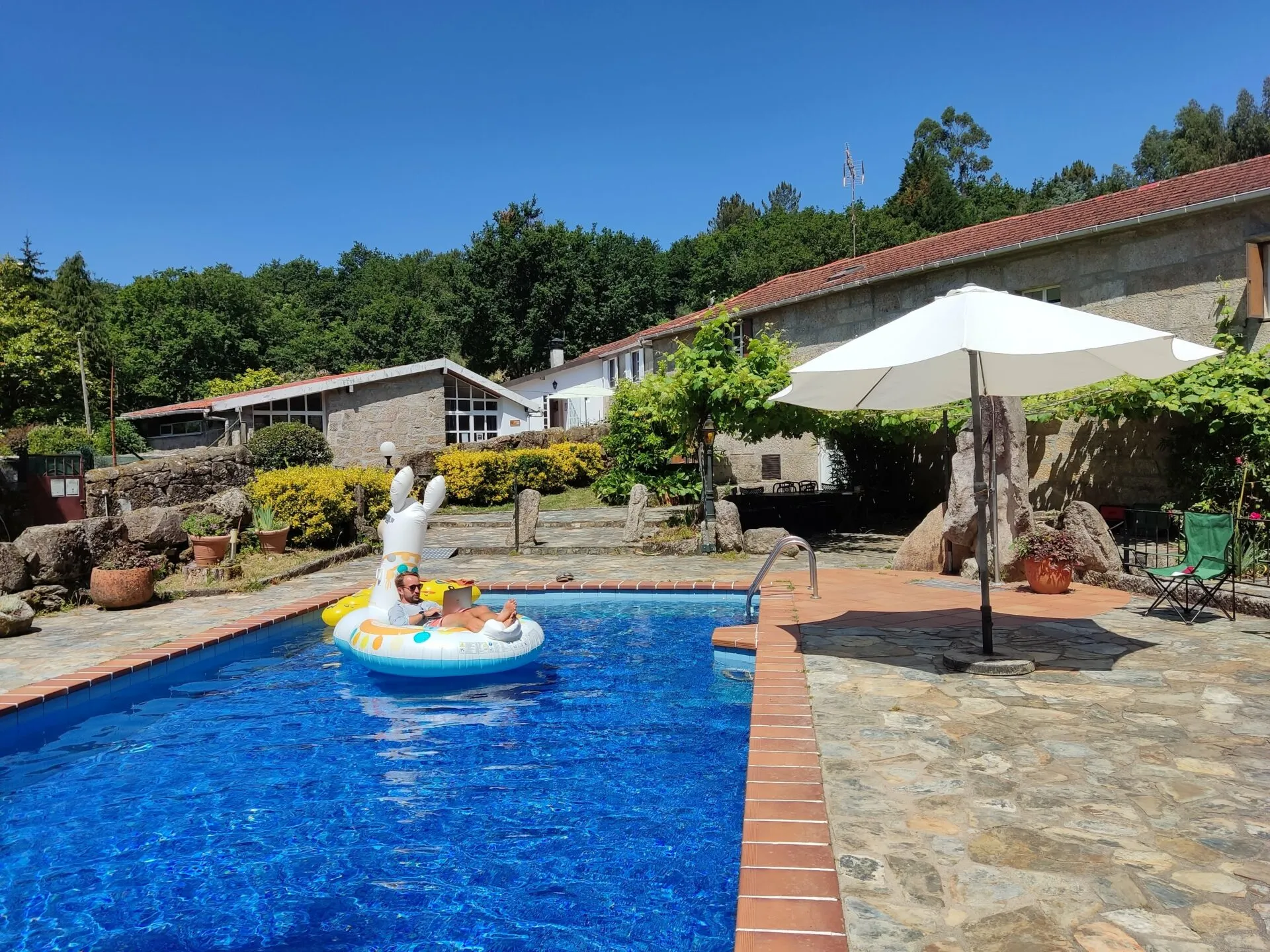 A person floating on an inflatable ring in a blue swimming pool surrounded by stone paving, with a parasol and low stone buildings behind