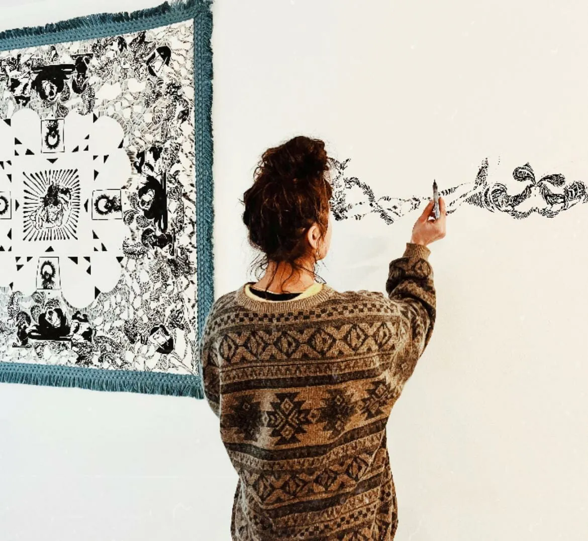 Woman seen from behind drawing an intricate black pattern straight onto a white wall beside a fringed tapestry of similar artwork