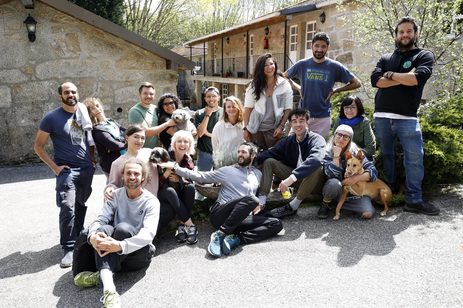 A group of about sixteen people posing on a gravel courtyard in front of a stone house, with two dogs among them