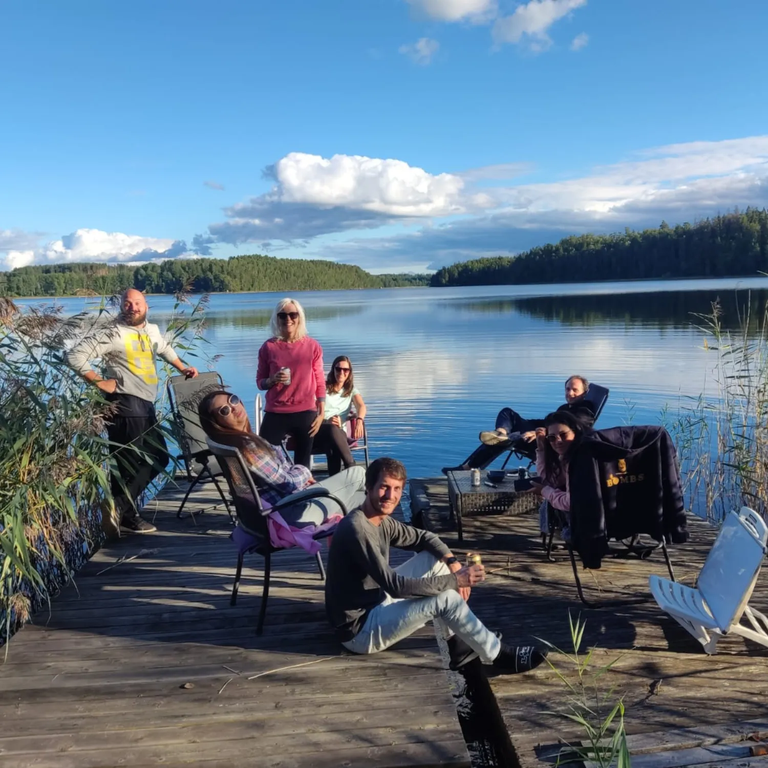 Seven people relaxing in folding chairs on a wooden jetty beside a calm forest-ringed lake under a blue sky