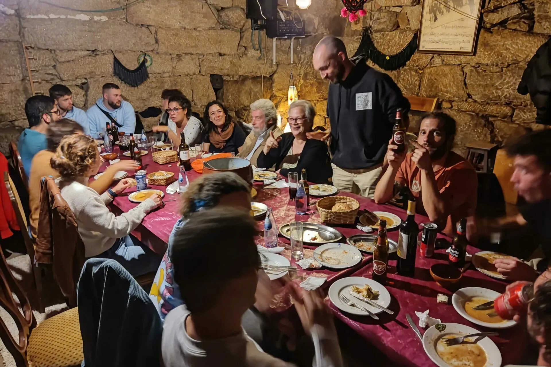 Long dinner table packed with people, plates and beer bottles in a stone-walled cellar, one man standing with a bottle raised