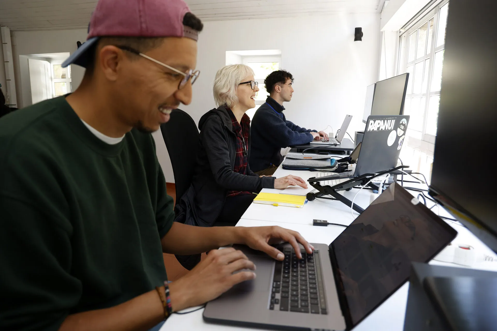 Three people typing on laptops along a long white desk in a bright, white-walled coworking room