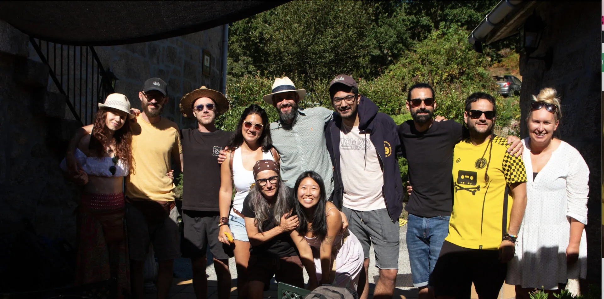 Eleven people in hats and sunglasses posing arm in arm in the shade beside a stone building