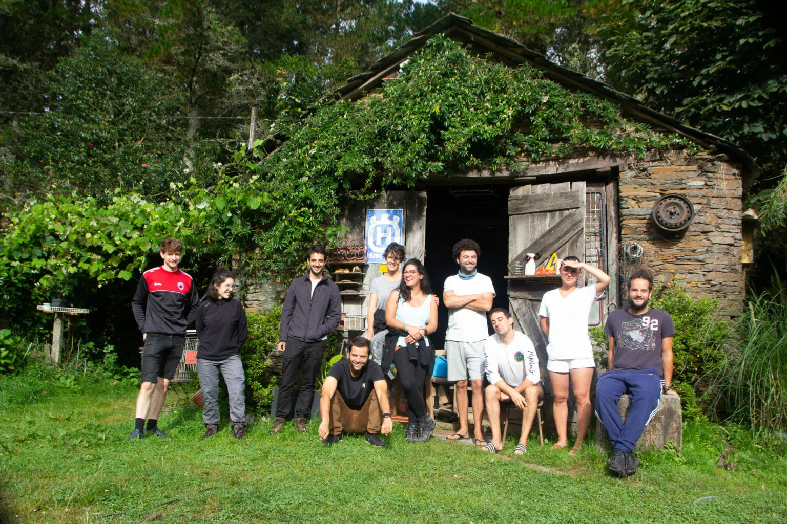 Ten young people posing on the grass in front of a vine-covered stone shed