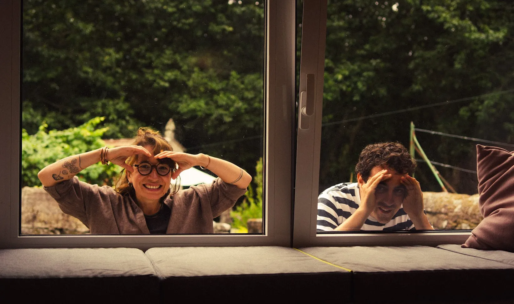 Two people peering in through a window from the garden, hands framing their faces, seen from a cushioned window seat inside