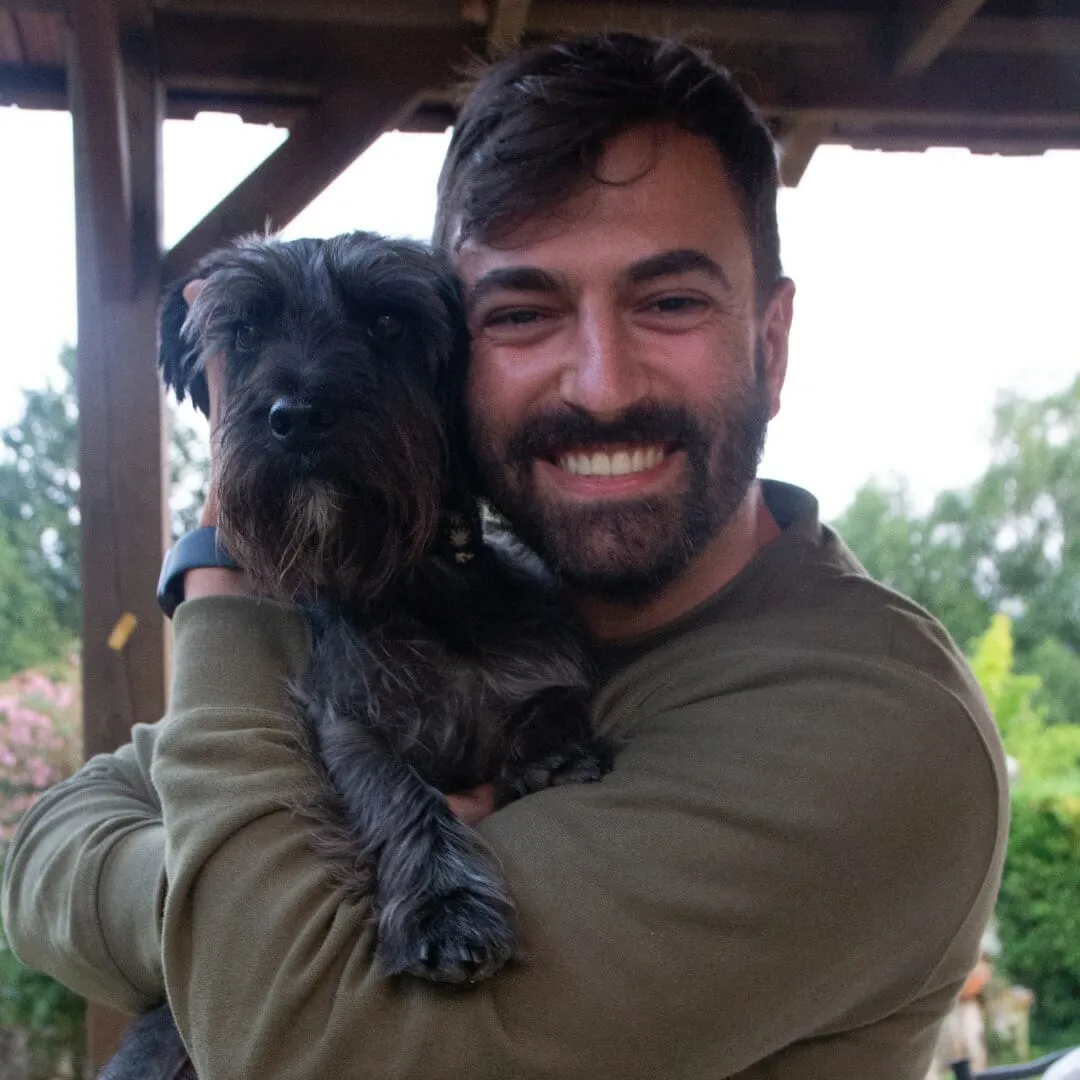 Smiling bearded man holding a small black wire-haired dog in his arms under a wooden porch