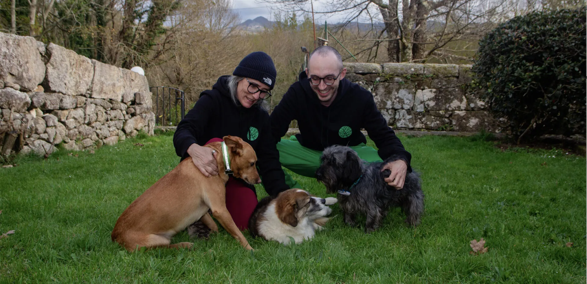 Two people in dark hoodies crouching on a lawn with three dogs in front of a stone wall