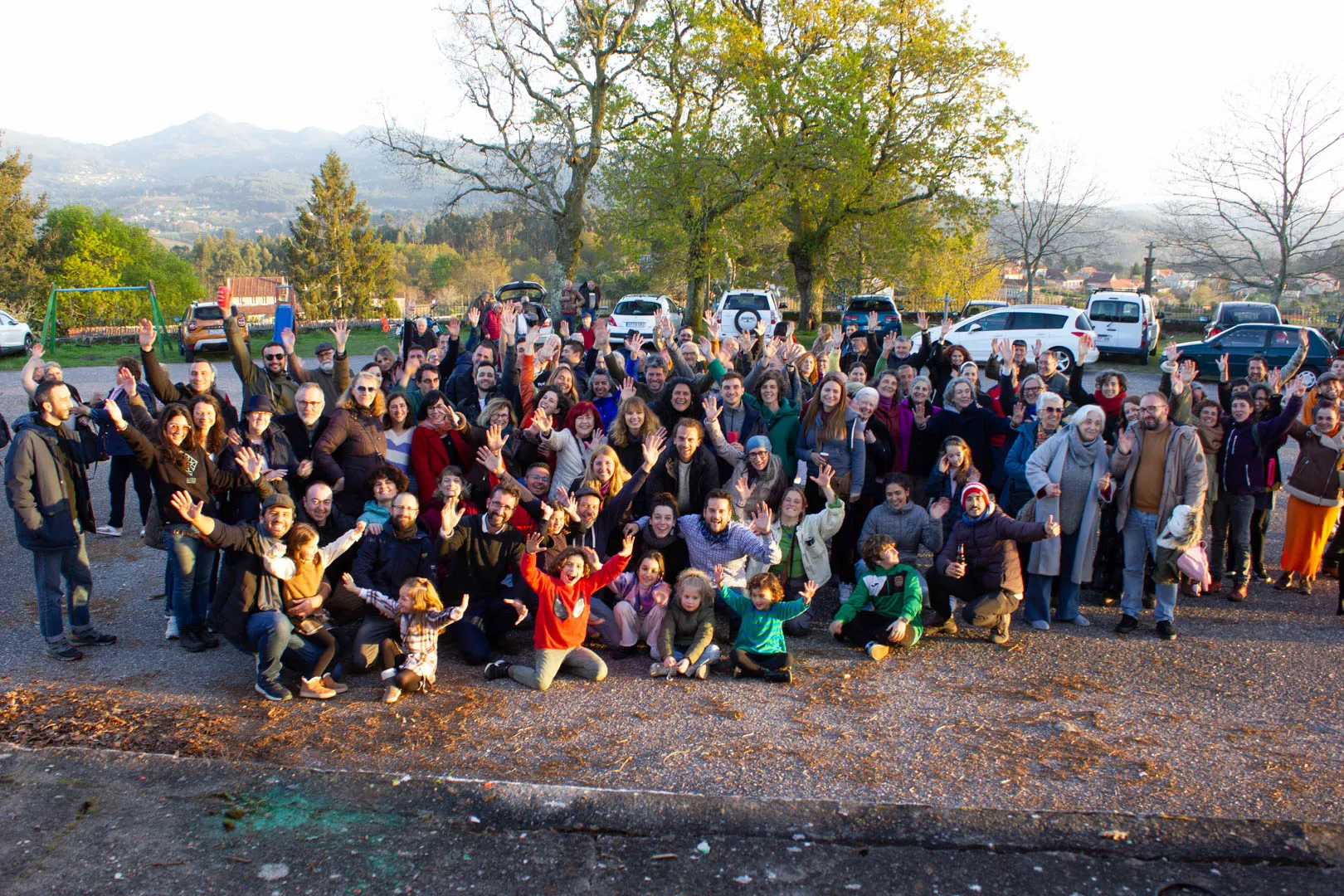 A very large crowd of adults and children gathered in a car park at sunset, arms raised, with mountains behind