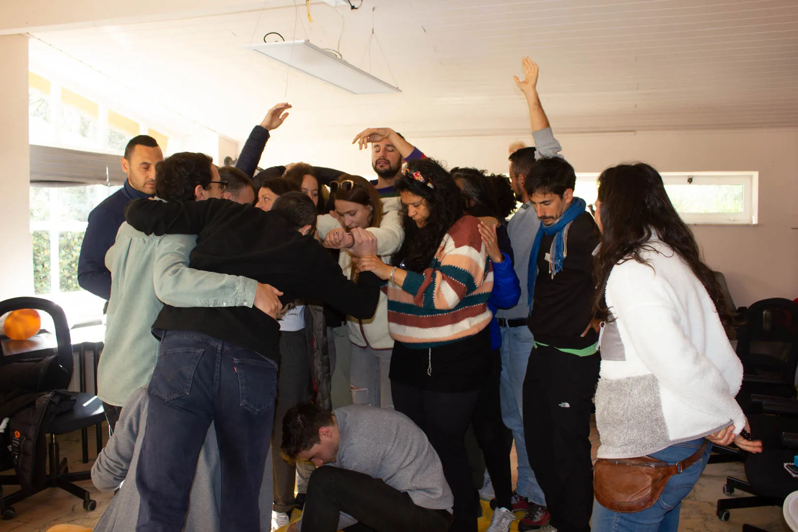 Workshop group huddled together with arms interlinked in a bright white room, a few hands raised above the tangle