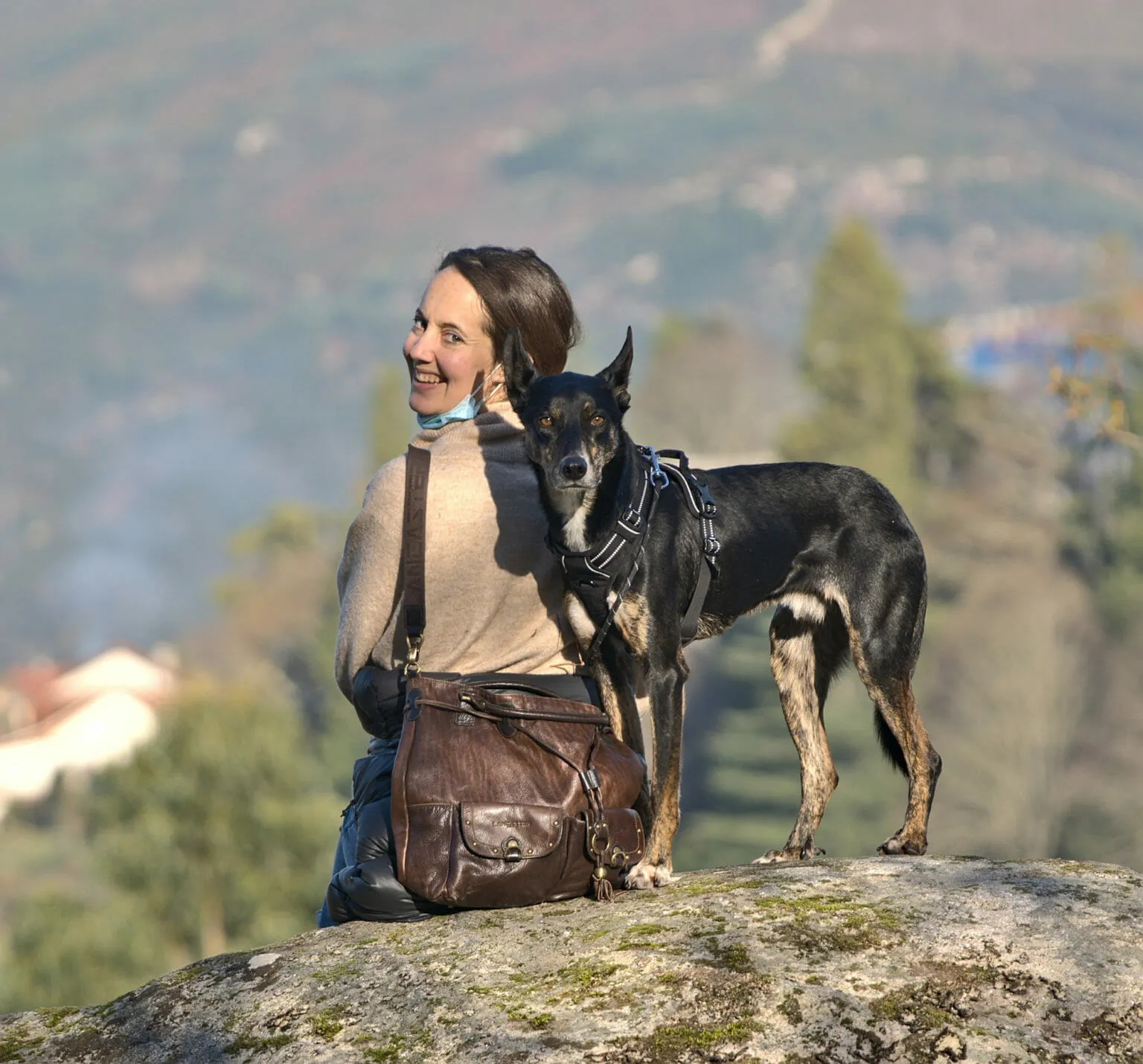 Smiling woman sitting on a mossy granite boulder beside a tall black-and-tan dog in a harness, valley behind