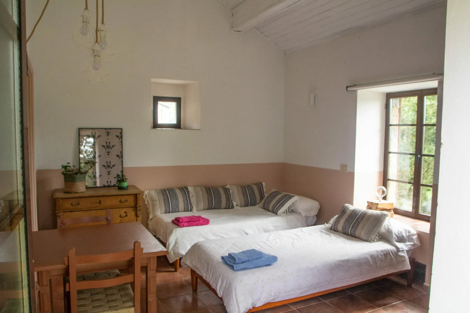 A bright whitewashed room with two single beds with striped cushions, a wooden chest of drawers and a window with shutters