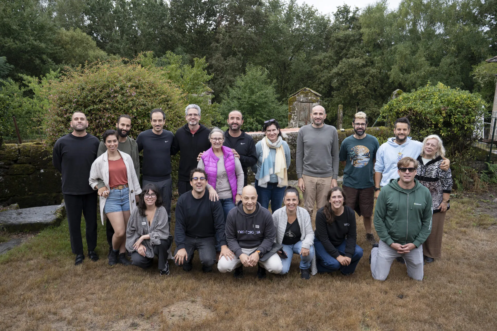 About twenty people posing together on dry grass in front of old stone walls and thick greenery
