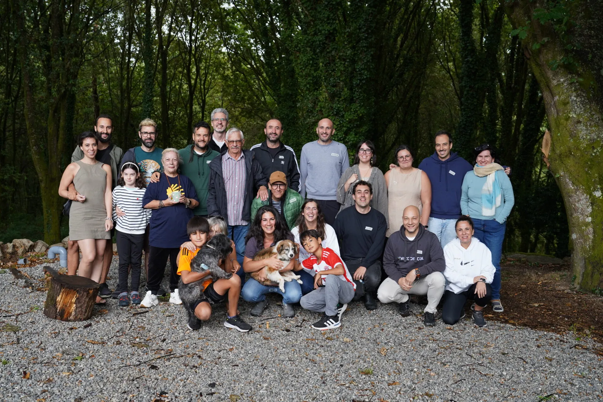 Group of about twenty-five adults and children with two dogs posing on gravel in front of dense green woodland
