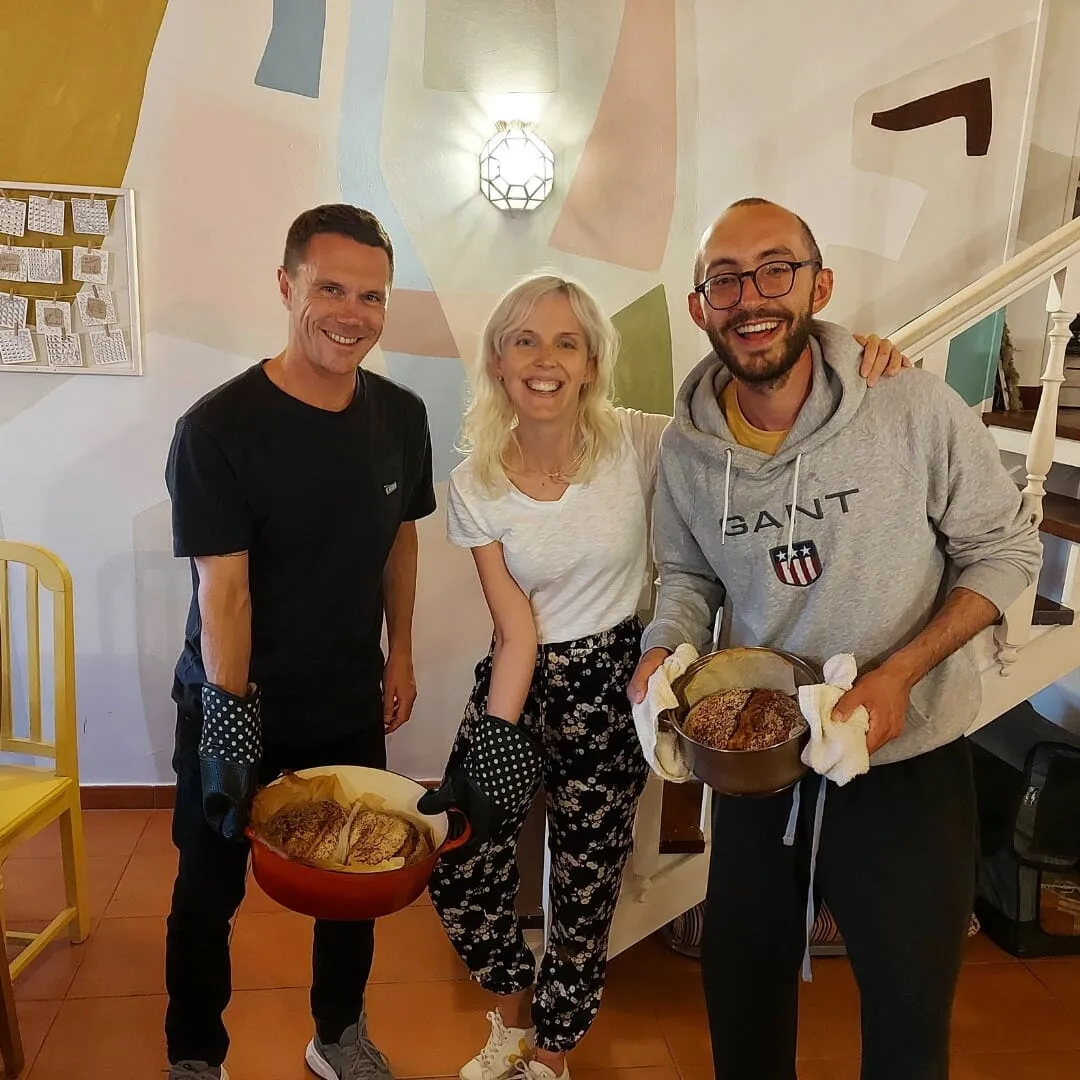 Three smiling colivers holding cast-iron pots of freshly baked bread in front of a pastel mural wall