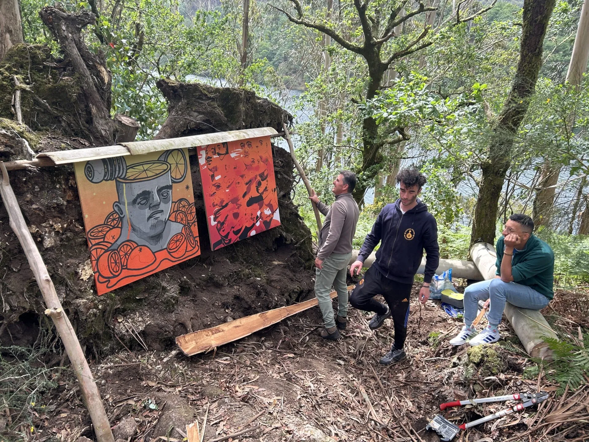 Two brightly painted panels mounted on an upturned tree root in the riverside forest, with three men beside them