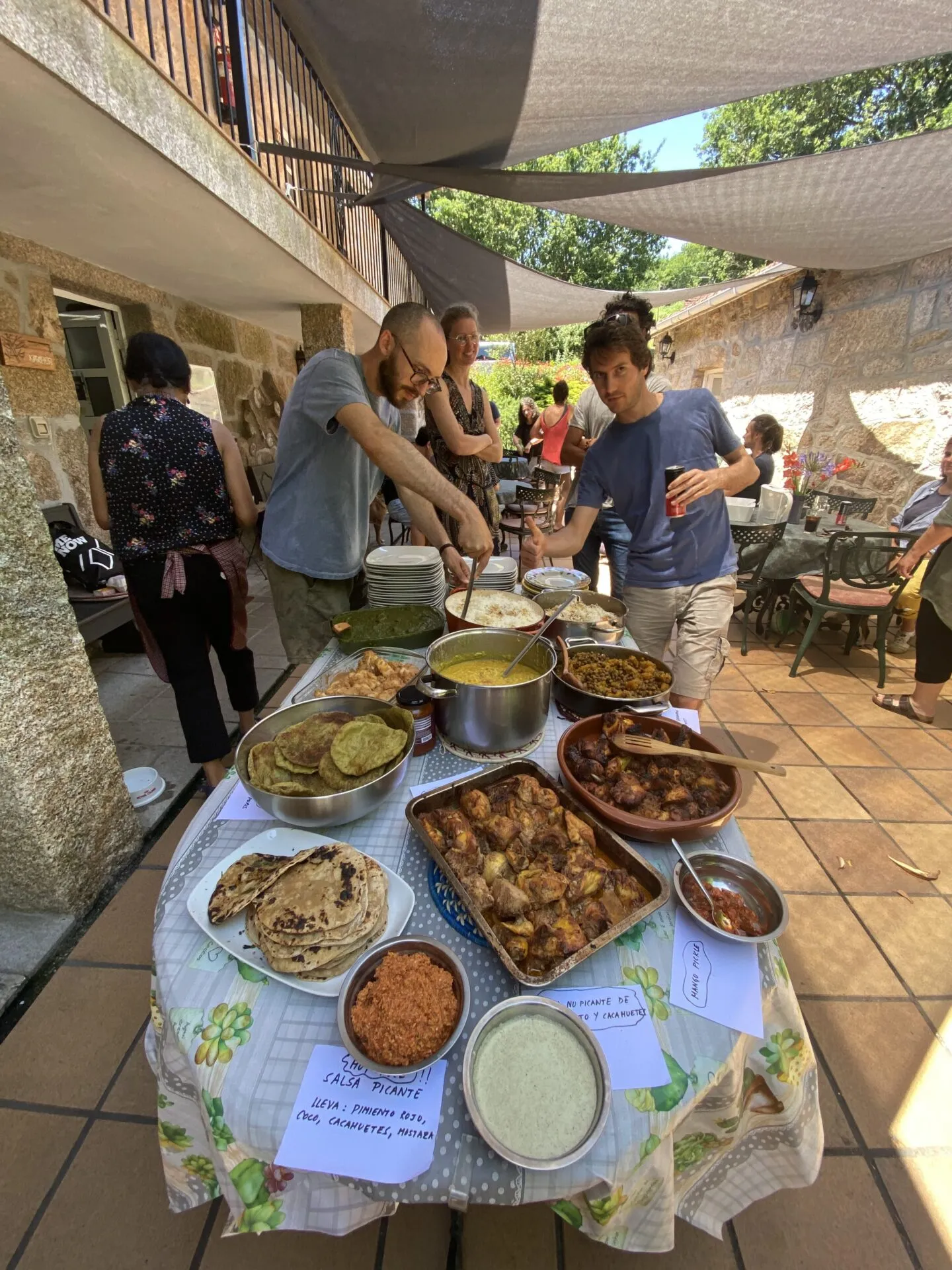 Round table on a shaded terrace loaded with curries, rice and flatbreads, with handwritten labels reading “salsa picante” and “mango pickle”
