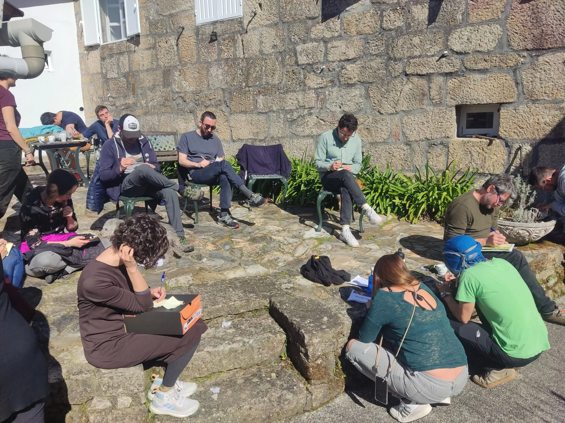 A dozen people scattered across a sunny stone courtyard, writing on notepads against a granite wall