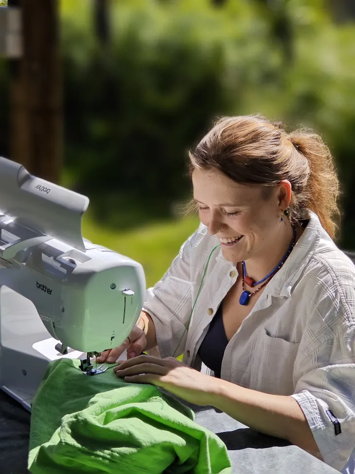 Woman smiling as she sews green fabric on a sewing machine set up outdoors against a leafy background