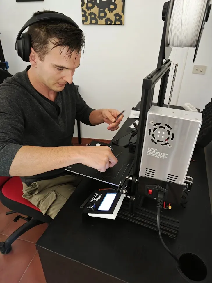 Man wearing headphones working at a desktop 3D printer loaded with a spool of white filament