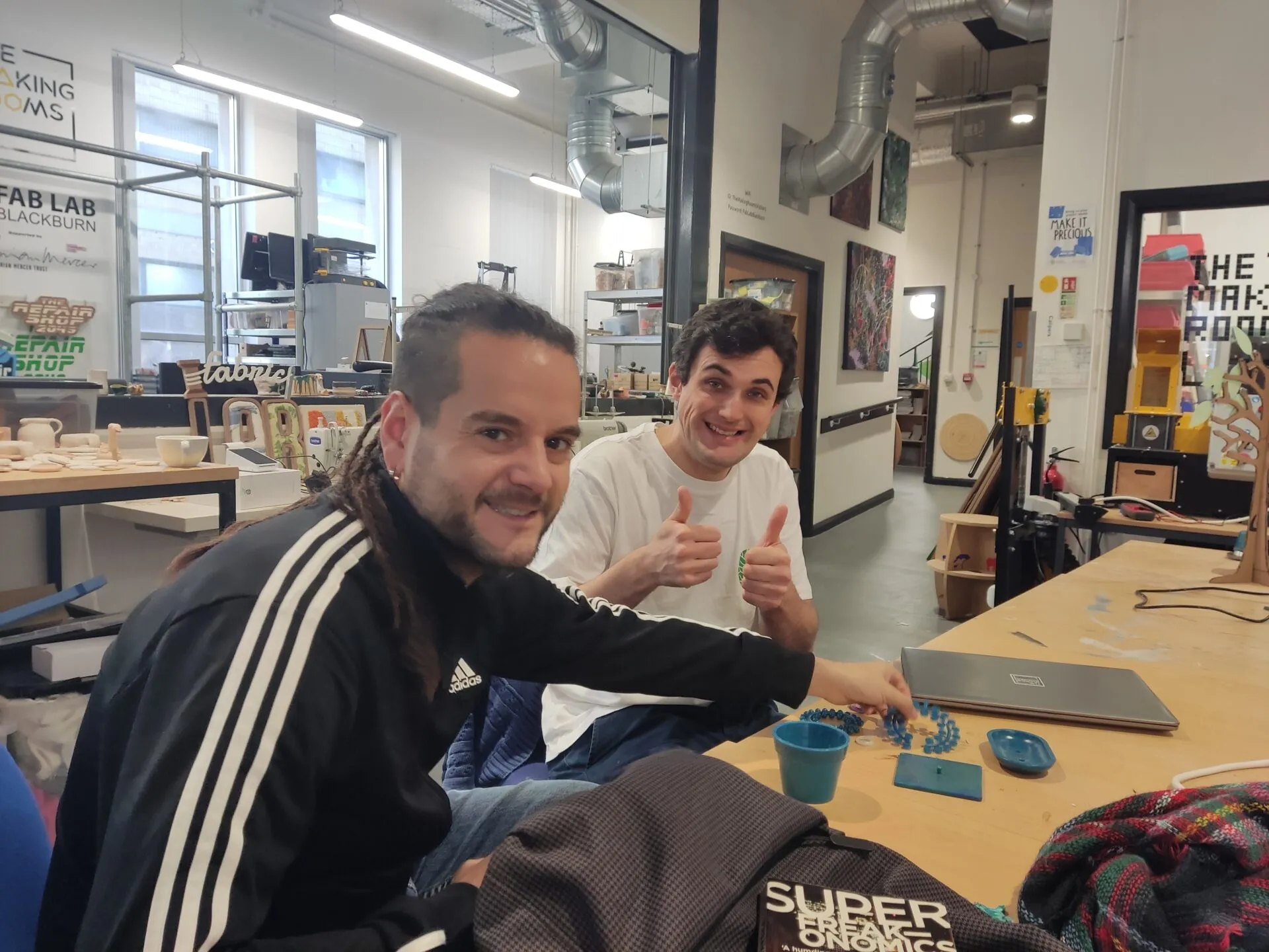 Two men smiling at a workbench with blue 3D-printed pieces in a workshop signed FAB LAB BLACKBURN and The Making Rooms