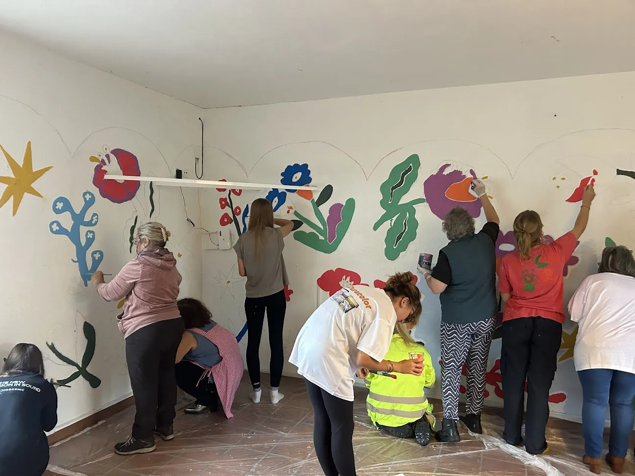 Around eight women painting a bright floral mural across the walls of a white room, the floor covered with plastic sheeting