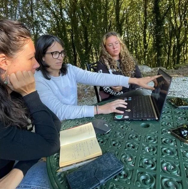 Three women around a garden table, one pointing at a laptop screen beside an open notebook