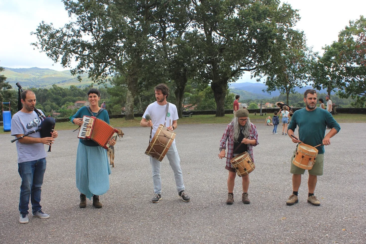 Five musicians playing bagpipes, accordion and traditional drums on a square with mountains behind