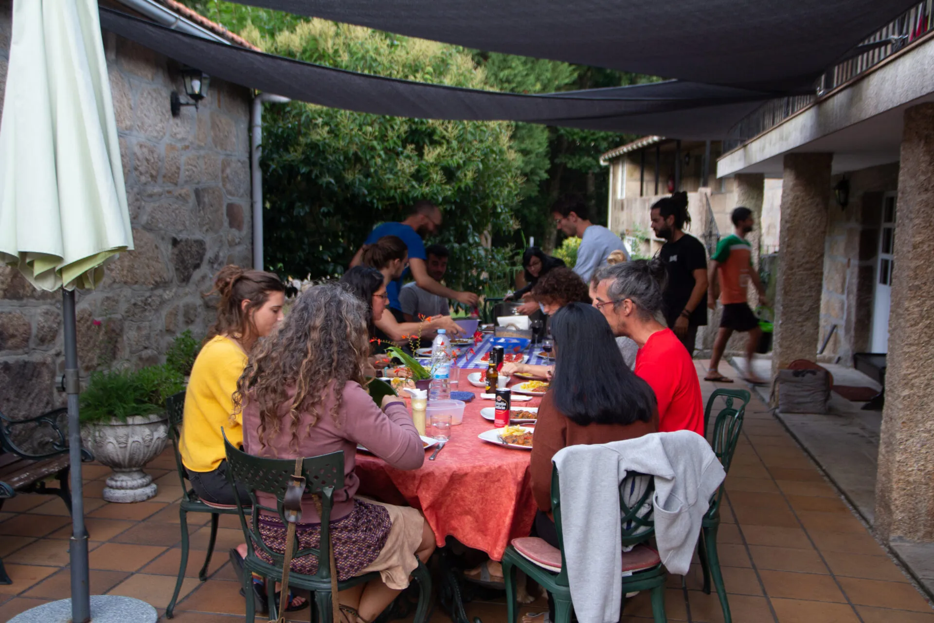 A dozen people eating together at a long table under a shade sail on the tiled terrace of the stone house