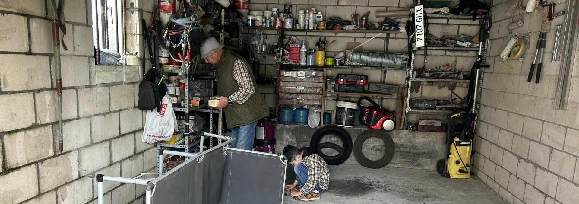 An older man working at a bench in a cluttered concrete-block garage workshop, while another person crouches on the floor
