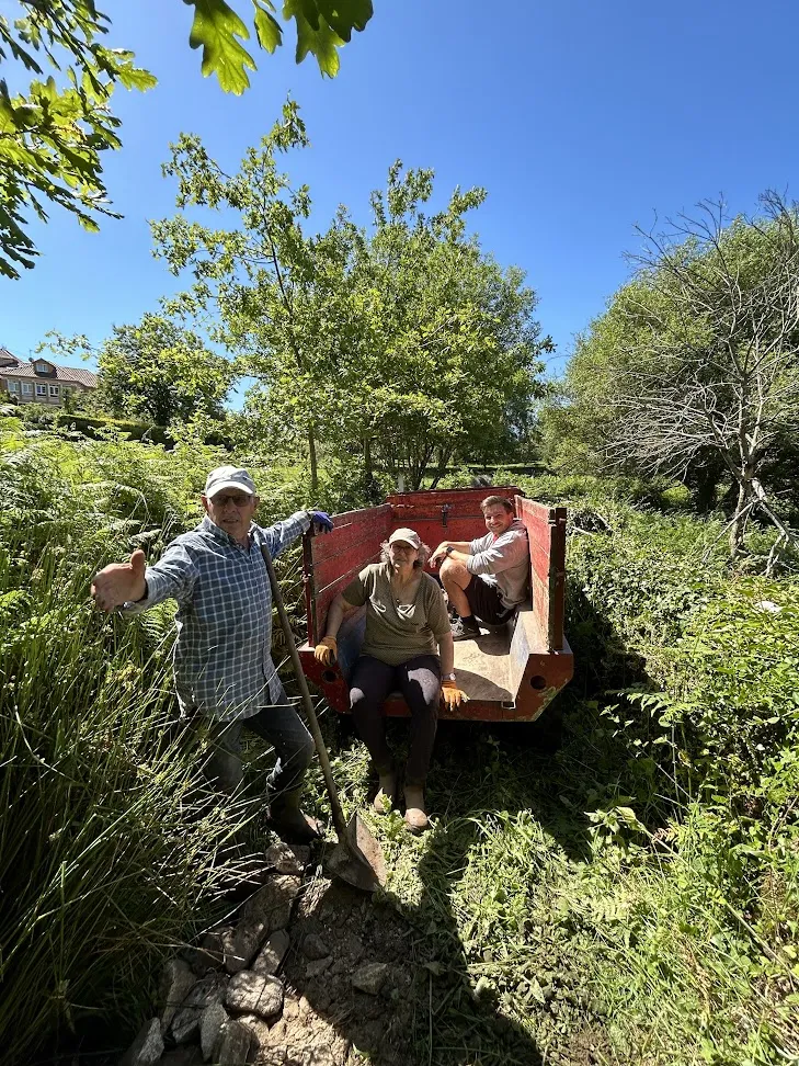 Man holding a shovel and two people sitting in a small red trailer on an overgrown green path