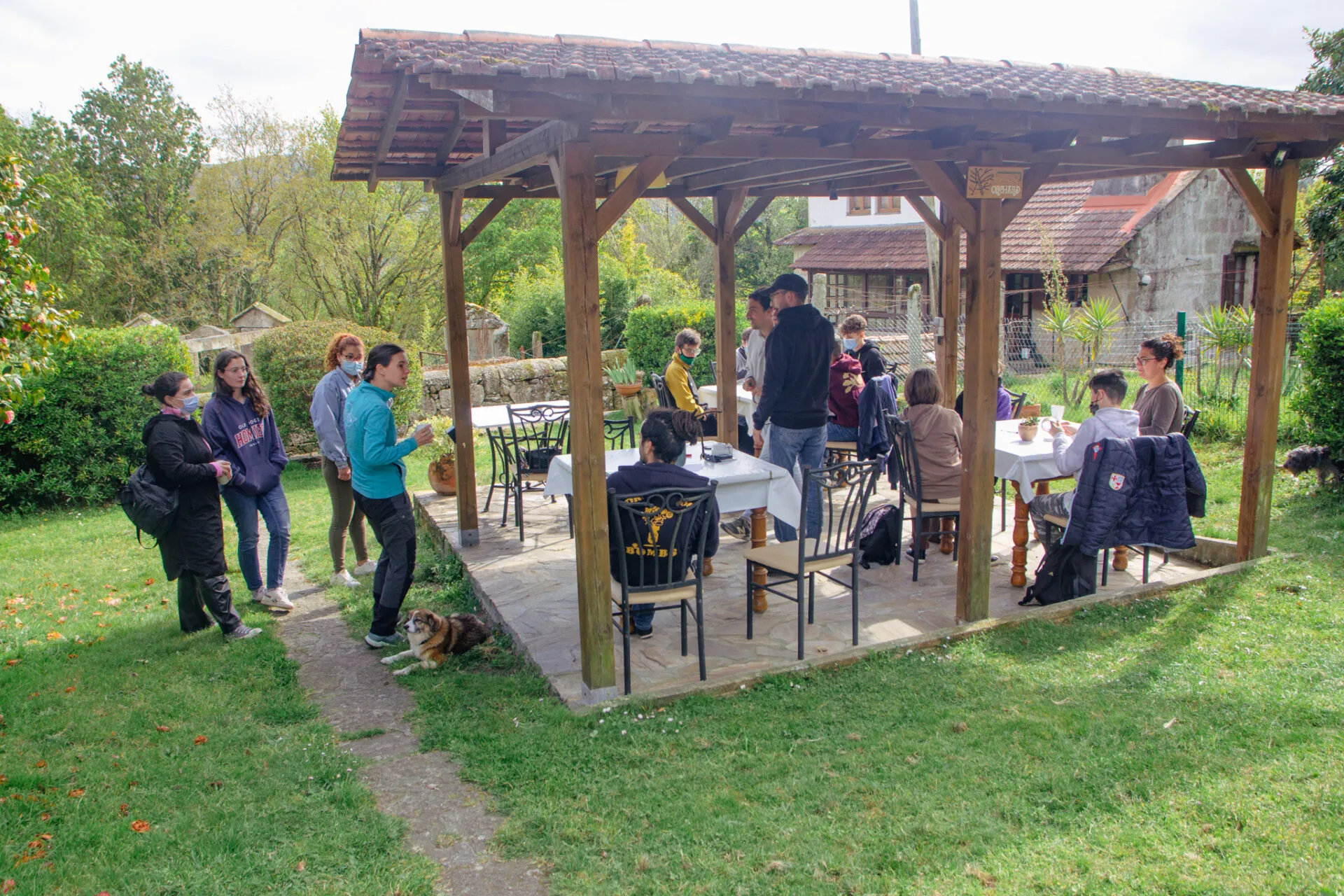 People with face masks gathered around tables under a wooden pavilion with a tiled roof in a green garden