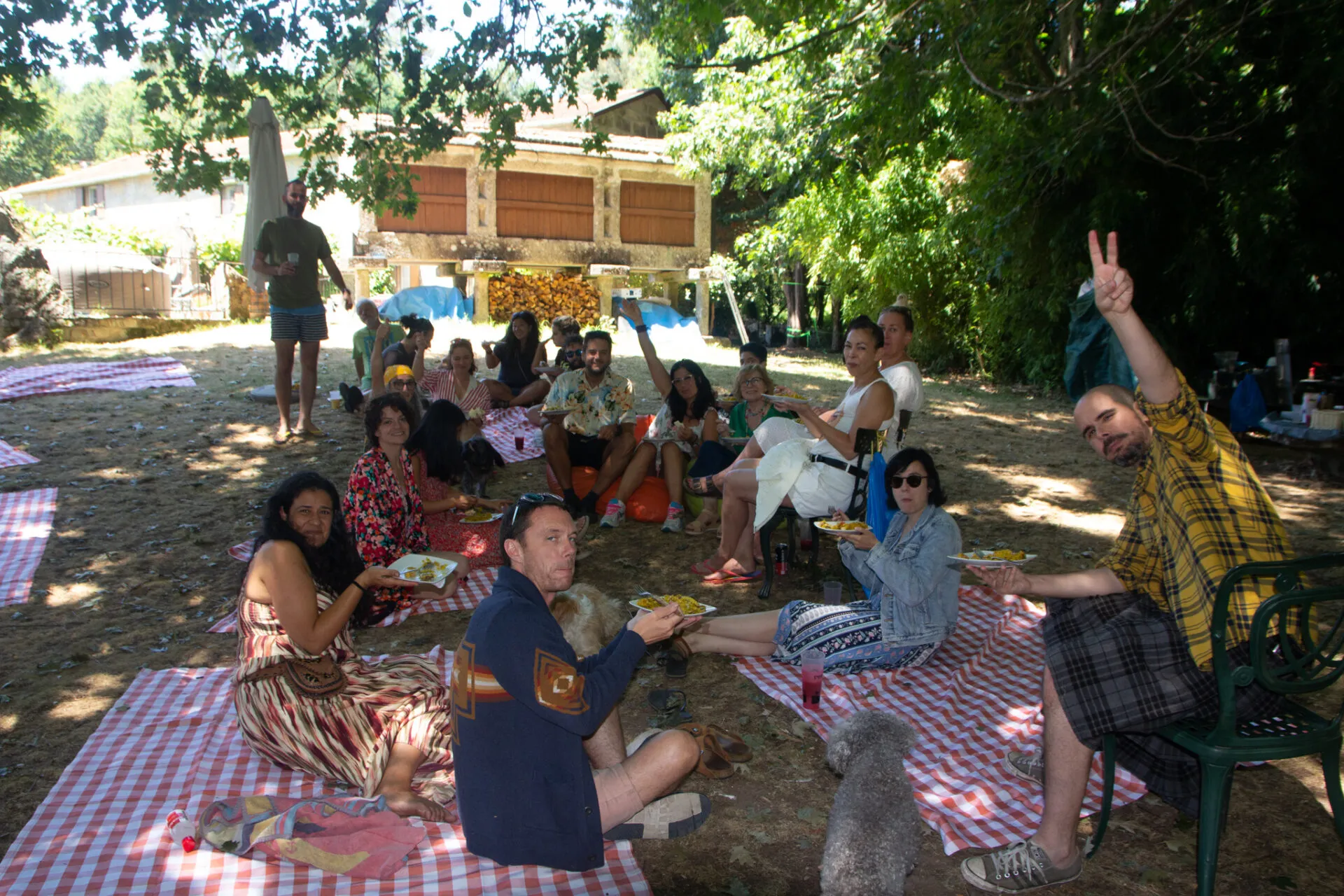 People sitting on checkered picnic blankets in the dappled shade of trees, with a stone building behind them