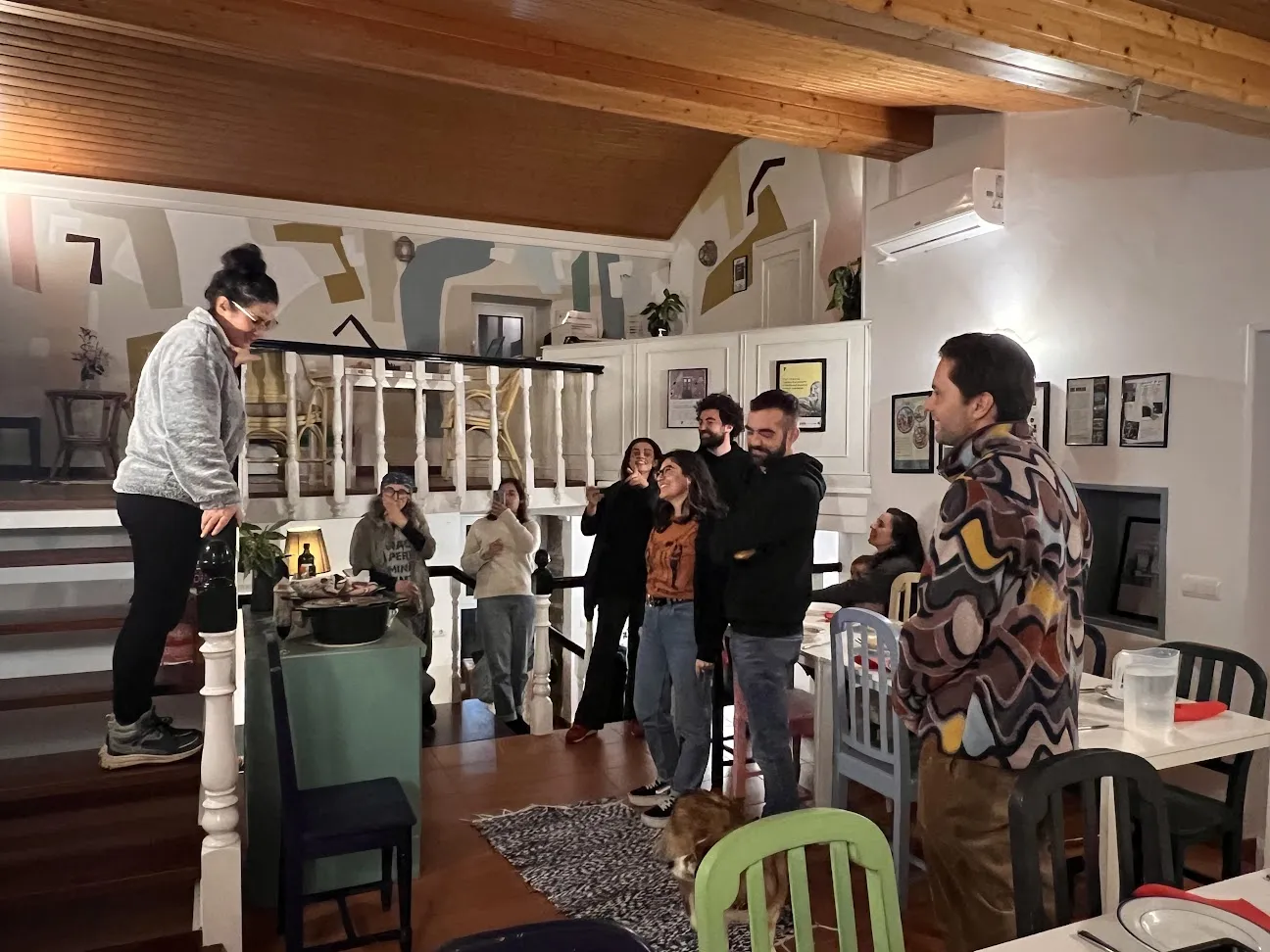 Woman standing on a raised step addressing a laughing group in a dining room with a painted mural and wooden ceiling