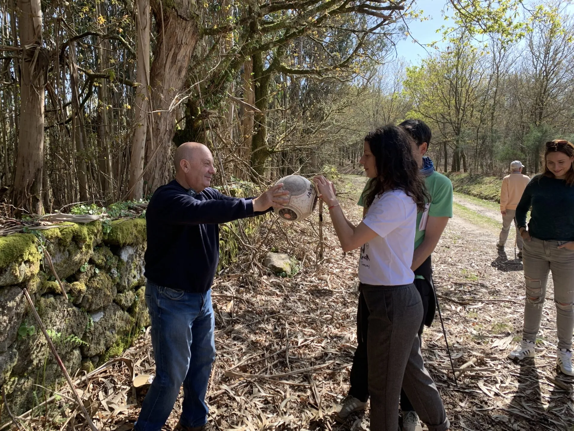 A man handing a round clay pot to a woman on a woodland path lined with a mossy stone wall