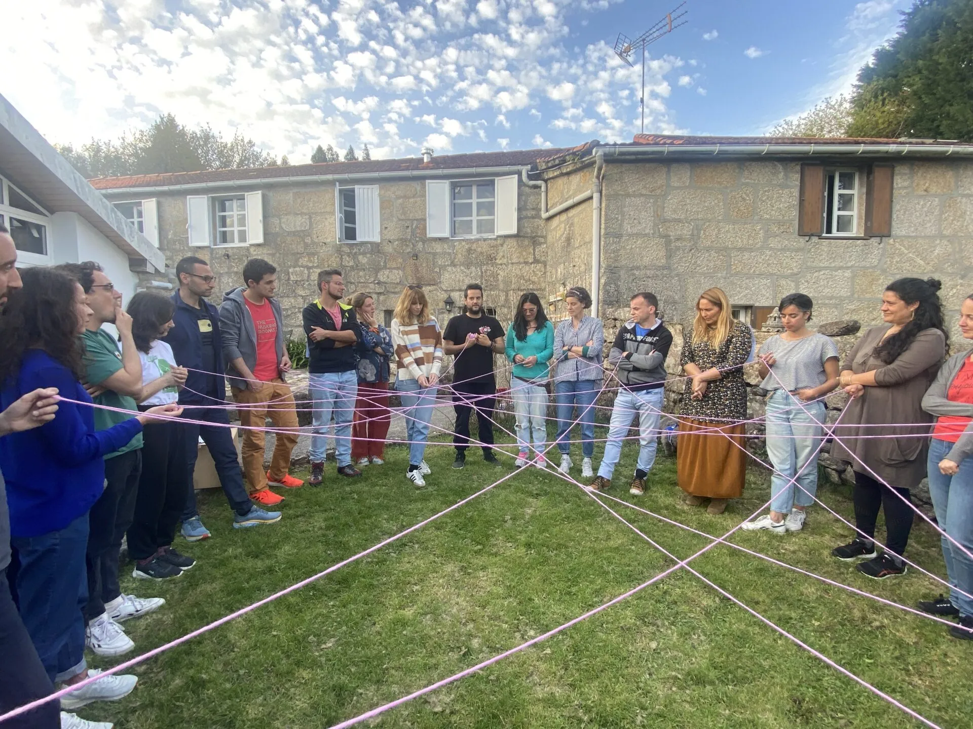 Around thirty people standing on a lawn holding a web of pink yarn stretched between them, in front of a stone house
