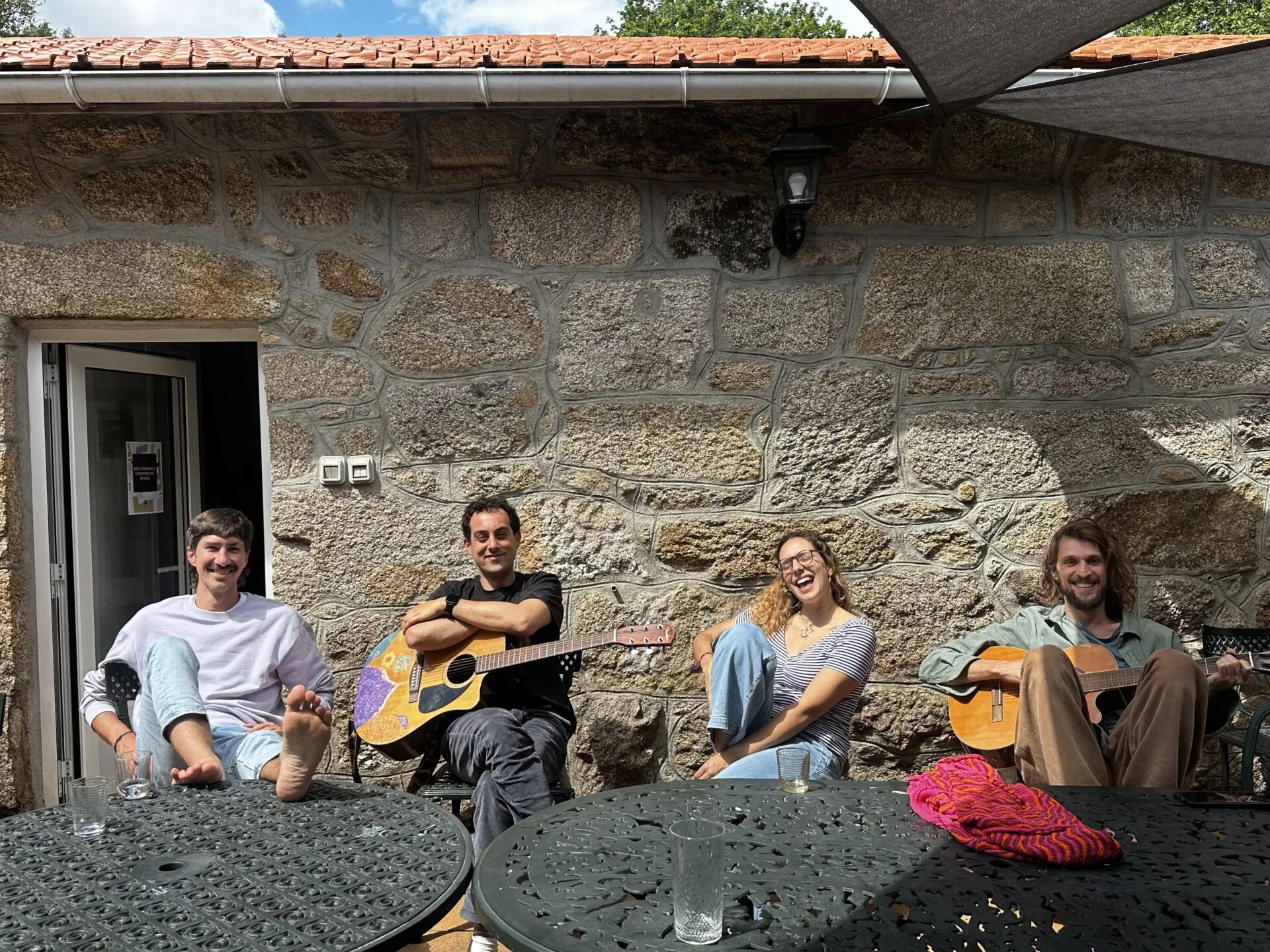 Four people relaxing against a granite wall on a patio, two of them playing acoustic guitars