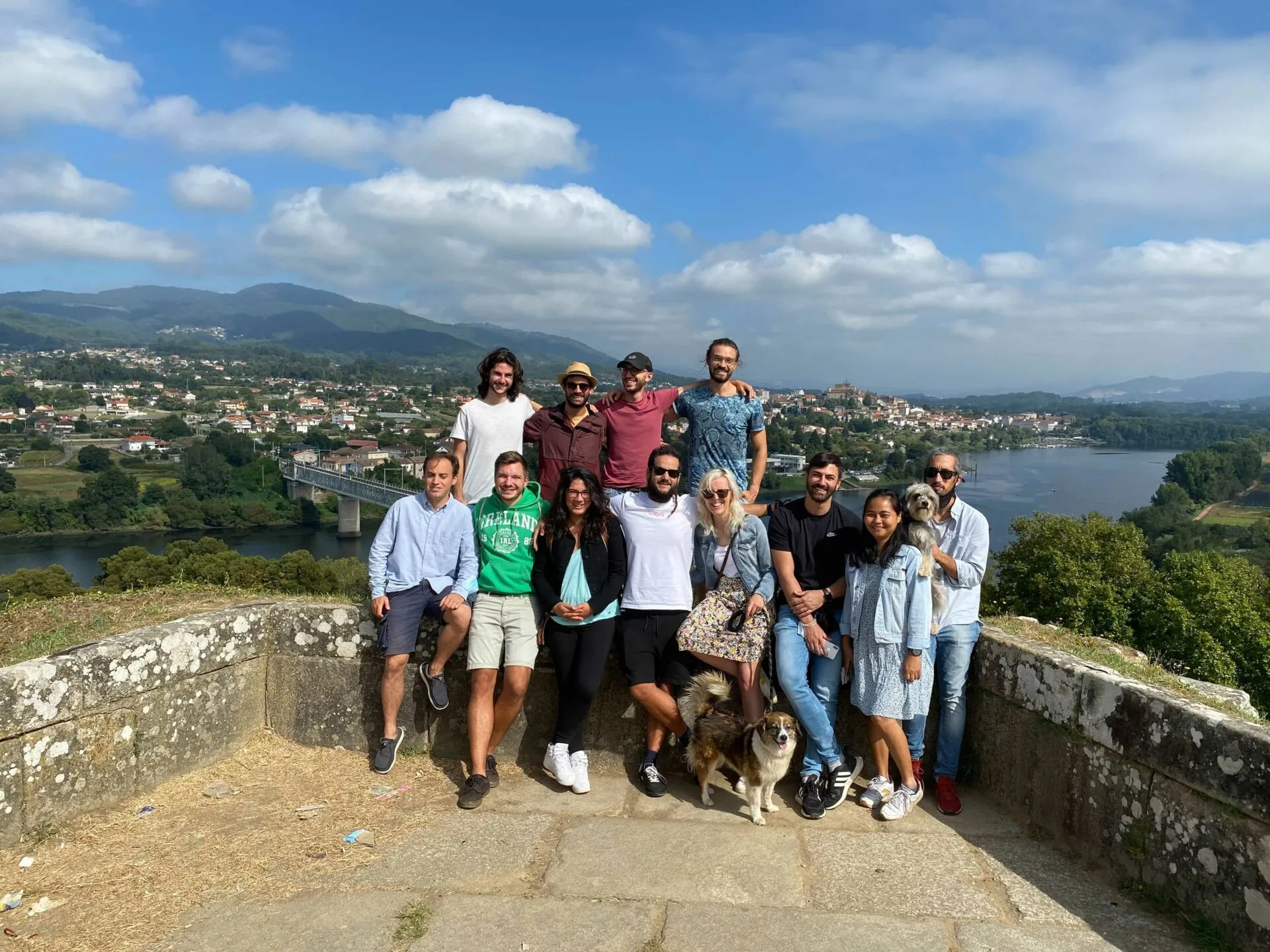 Eleven people and two dogs posing on a stone lookout wall with a river valley and a town behind them