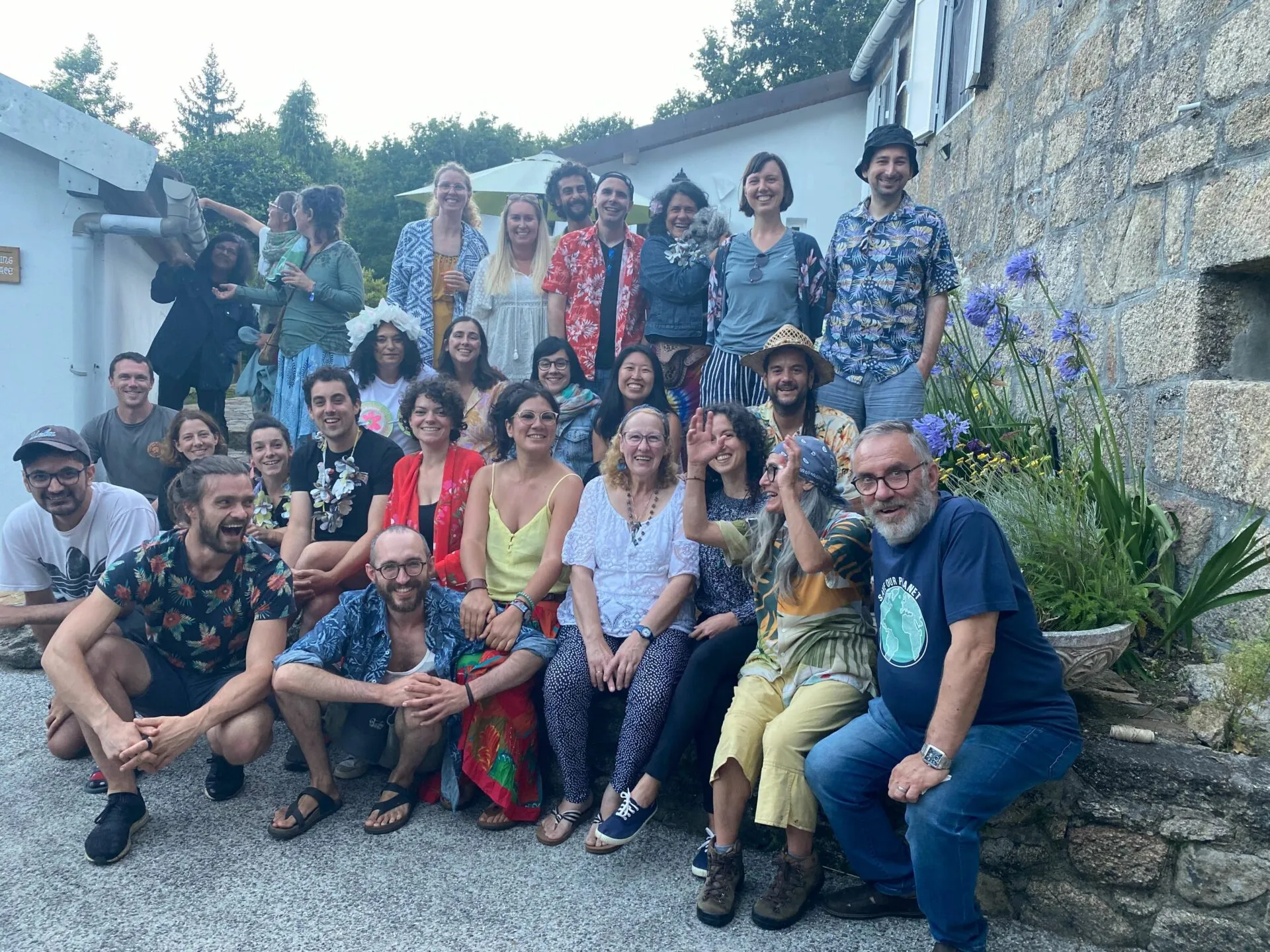 Around forty people in colourful summer clothes posing in a courtyard beside a stone house and blue agapanthus flowers