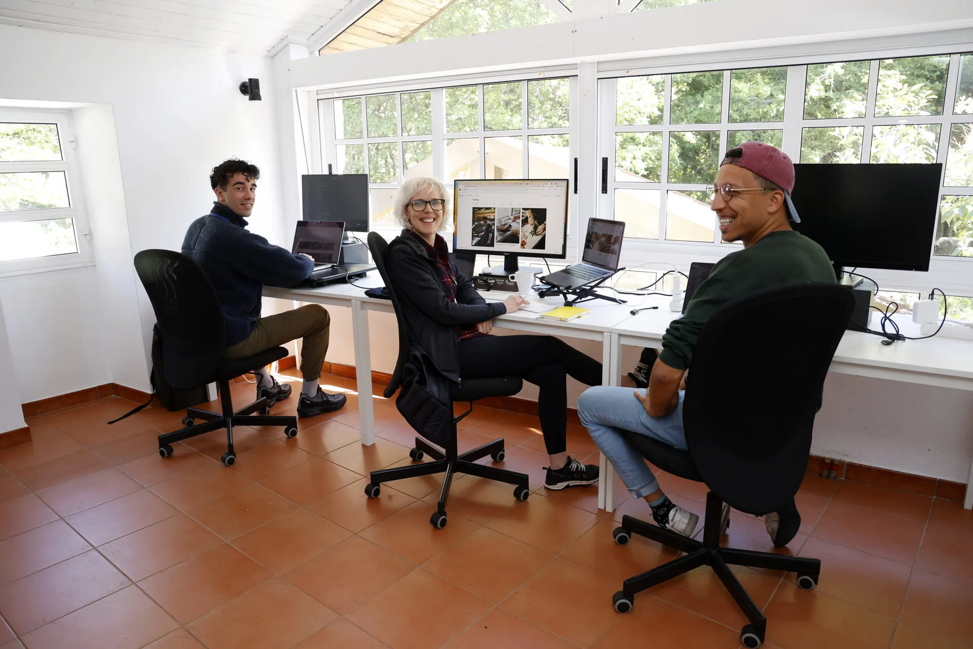 Three colivers at their desks in the glass-walled coworking room, turning to the camera and laughing, oak trees outside the windows